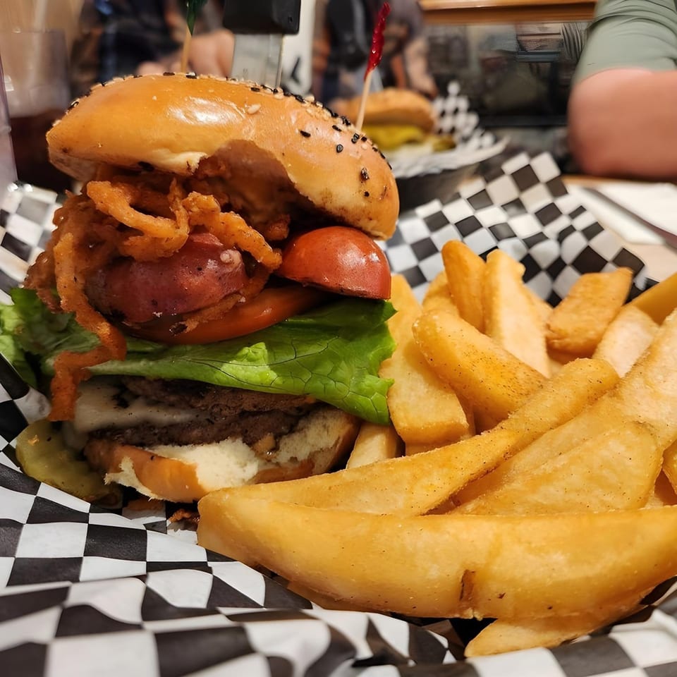 A Woody's Classic Grill burger with onion rings, tomato, lettuce, and fries in a checkered basket.
