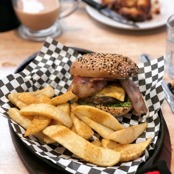 A bacon cheeseburger with sesame seed bun and a side of seasoned potato wedges in a black basket.